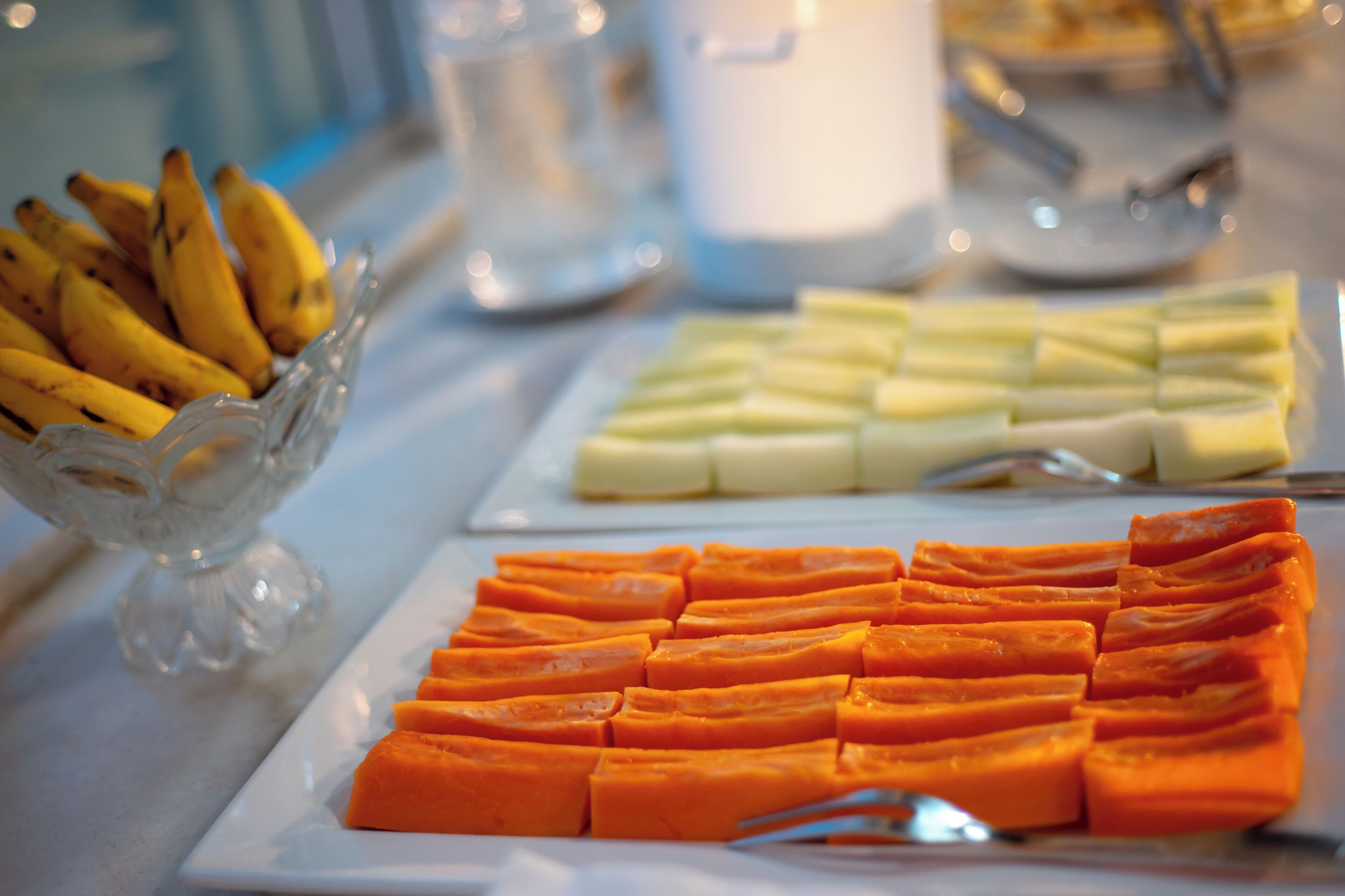 A buffet table with sliced fruits and a glass bowl of bananas.