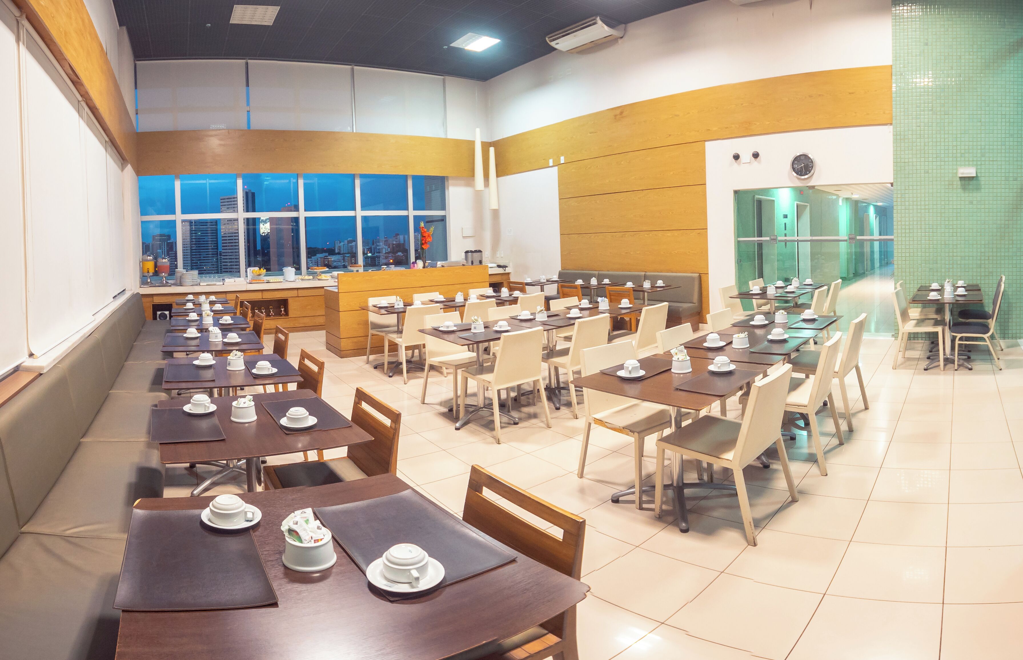Un vaste espace de salle à manger avec plusieurs tables et chaises, de grandes fenêtres offrant une vue sur la ville et un coin cuisine visible au fond.