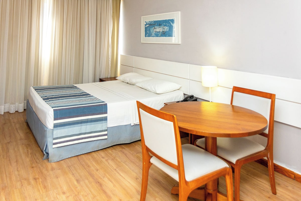 A hotel room with a bed, a bedside table with a lamp, a wooden chair, and a small table.
