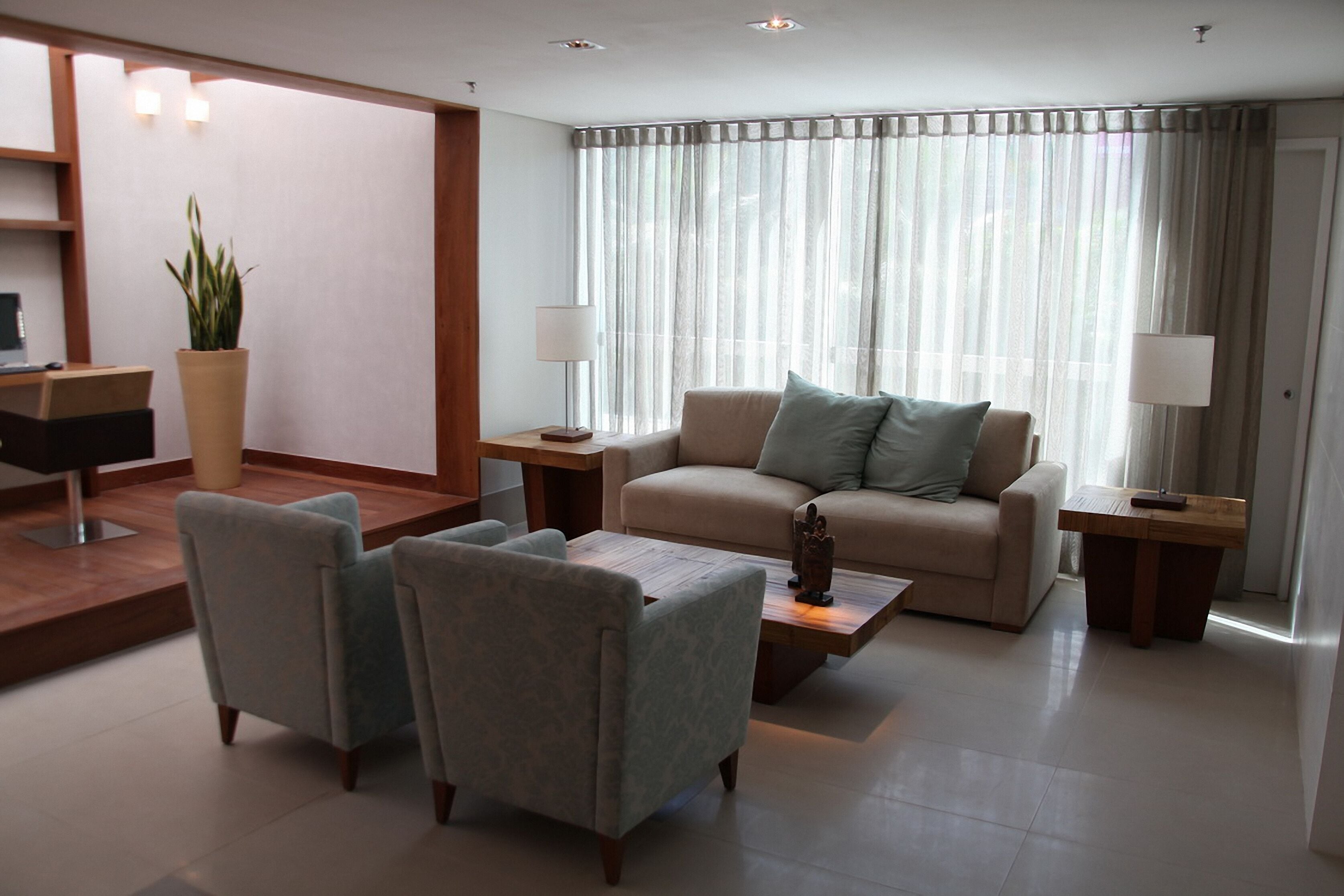 Un salon moderne comprenant un canapé, deux fauteuils, une table basse et une table d’appoint avec une lampe. On y trouve également une plante en pot et un bureau avec une chaise, placés dans un coin de la pièce.