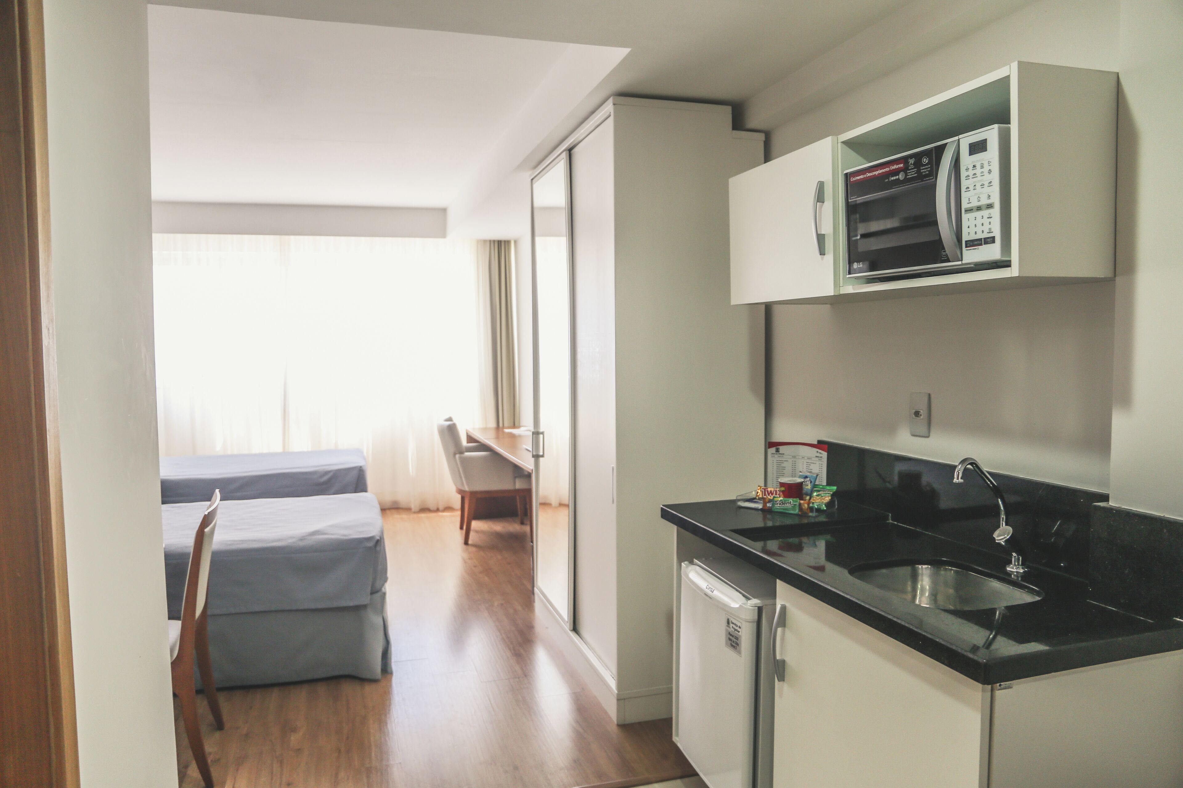 A compact kitchen with a microwave, a sink, and a small refrigerator. There is a dining area with a table and chairs visible through an open door.