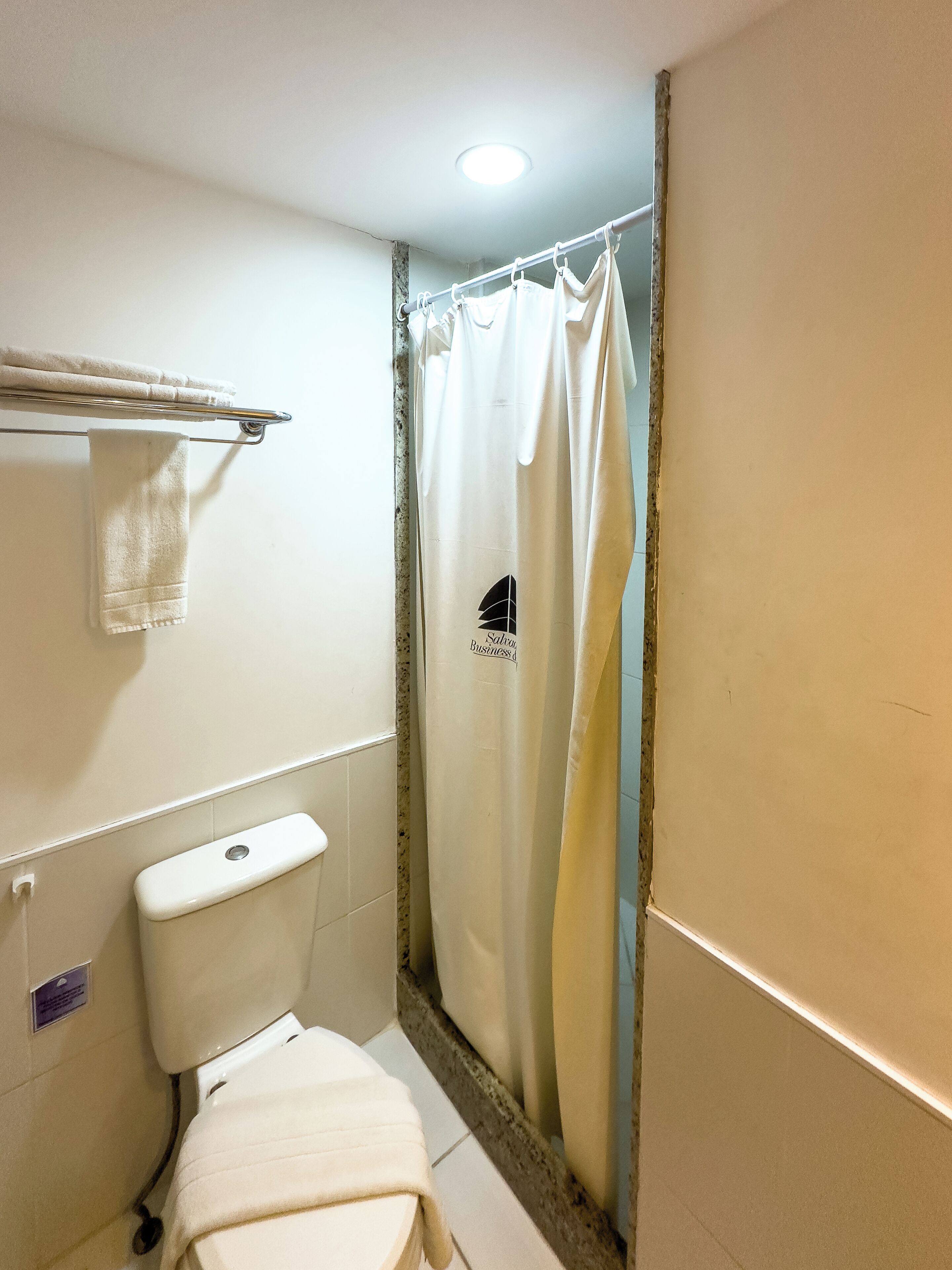 A bathroom with a toilet, a towel rack with towels, a shower curtain, and a visible sign on the wall.
