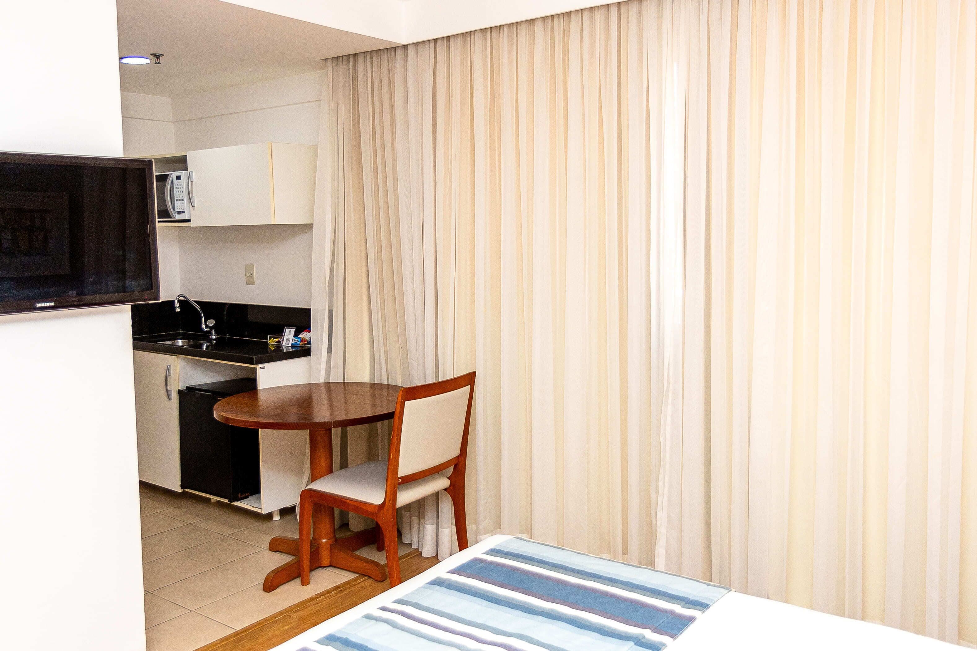 Una habitación de hotel compacta con kitchenette, una mesa de comedor y una silla, y un gran ventanal con cortinas.