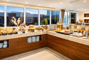 A modern kitchen with a breakfast buffet, wooden cabinets, and large windows offering a view of the cityscape.