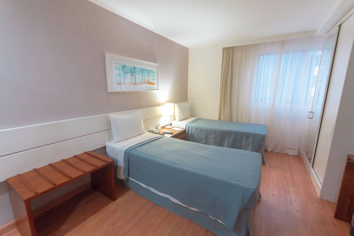 A hotel room with two beds, a bench, a nightstand with a lamp, a painting on the wall, and a large window with curtains.