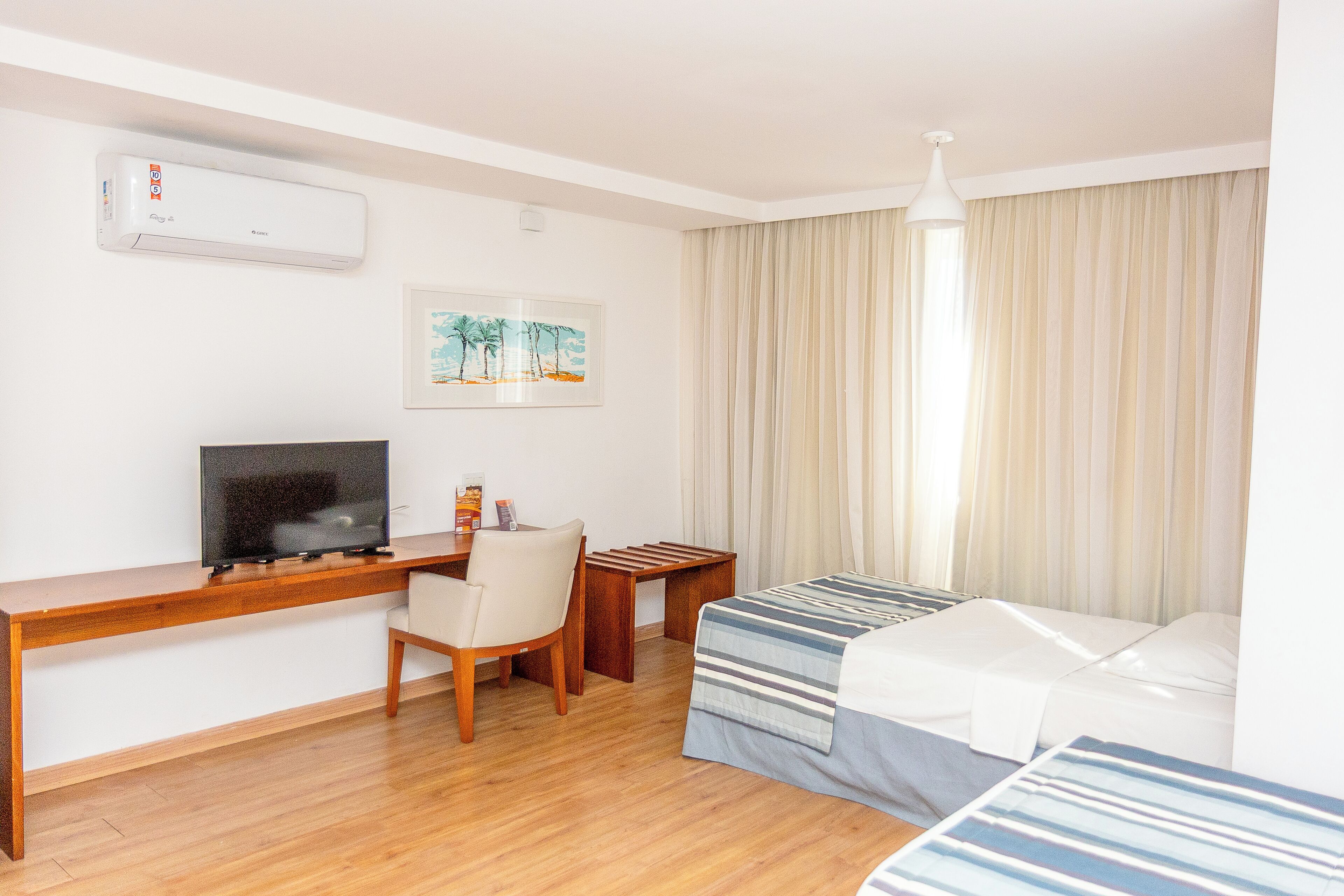 A hotel room with a wooden desk, a TV, a bed with striped bedding, a chair, a small table, and a window with curtains.