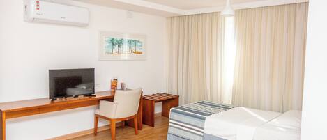 A hotel room with a wooden desk, a TV, a bed with striped bedding, a chair, a small table, and a window with curtains.