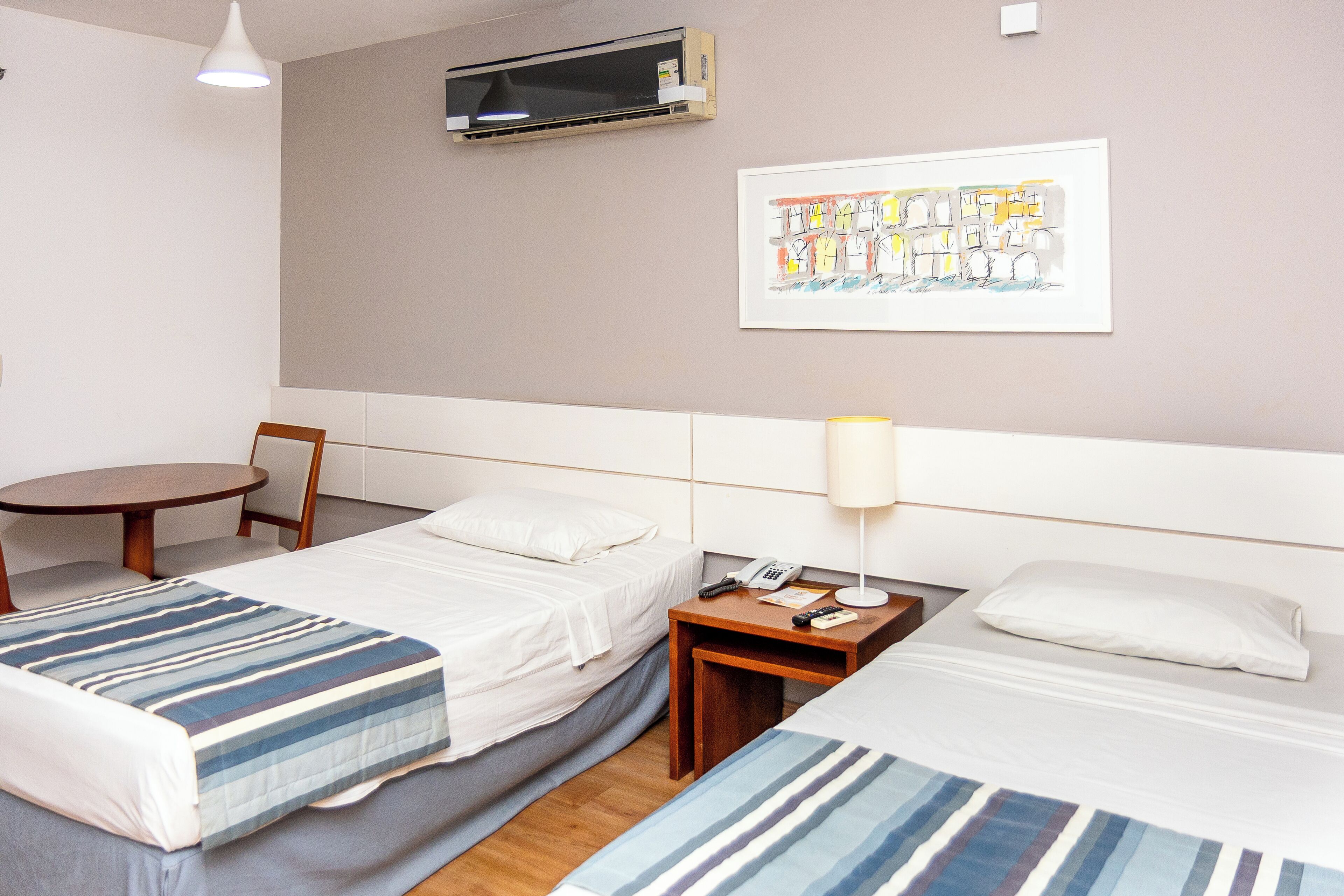 A hotel room with two beds, a small table, a chair, a lamp, an air conditioning unit, and a framed artwork on the wall.