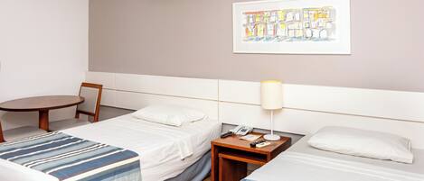 A hotel room with two beds, a small table, a chair, a lamp, an air conditioning unit, and a framed artwork on the wall.