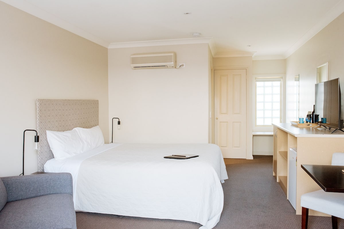 A hotel room with a bed, a desk, a chair, a small table, and a TV.