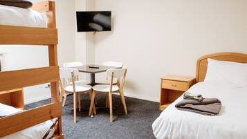 A hotel room with bunk beds, a small table with chairs, a TV mounted on the wall, and a bedside table with a book and towels.