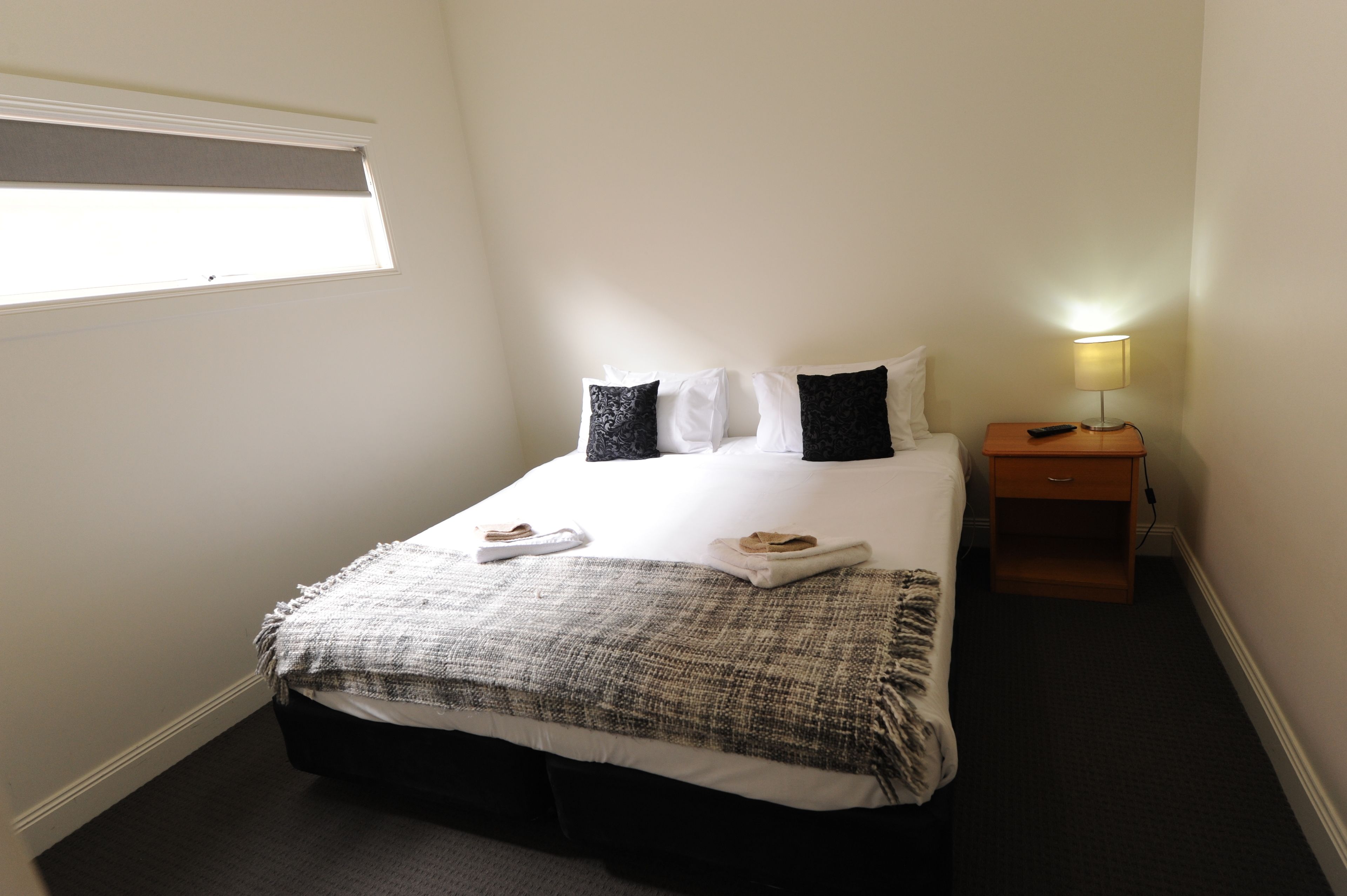 A neatly made bed with white and black pillows, a grey and white blanket, and two folded towels on the bed. There is a bedside table with a lamp and a remote control on it. The room has white walls and a window with blinds.