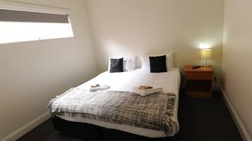 A neatly made bed with white and black pillows, a grey and white blanket, and two folded towels on the bed. There is a bedside table with a lamp and a remote control on it. The room has white walls and a window with blinds.