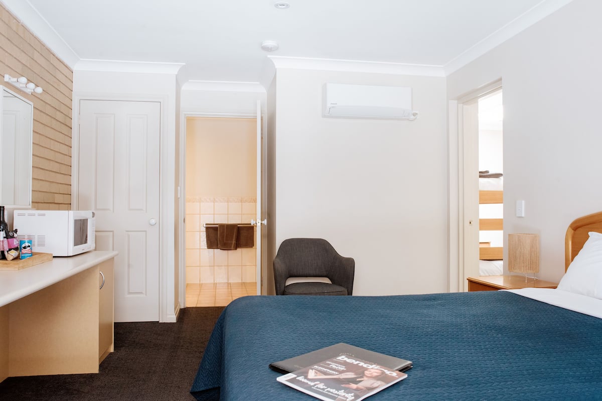 A hotel room with a bed, a desk with a microwave, a chair, and a bedside table with a lamp.