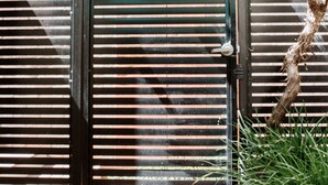 A closed metal gate with horizontal slats, a brick wall in the background, and a vine growing over the gate. - Alexandra Place (Bendigo)
