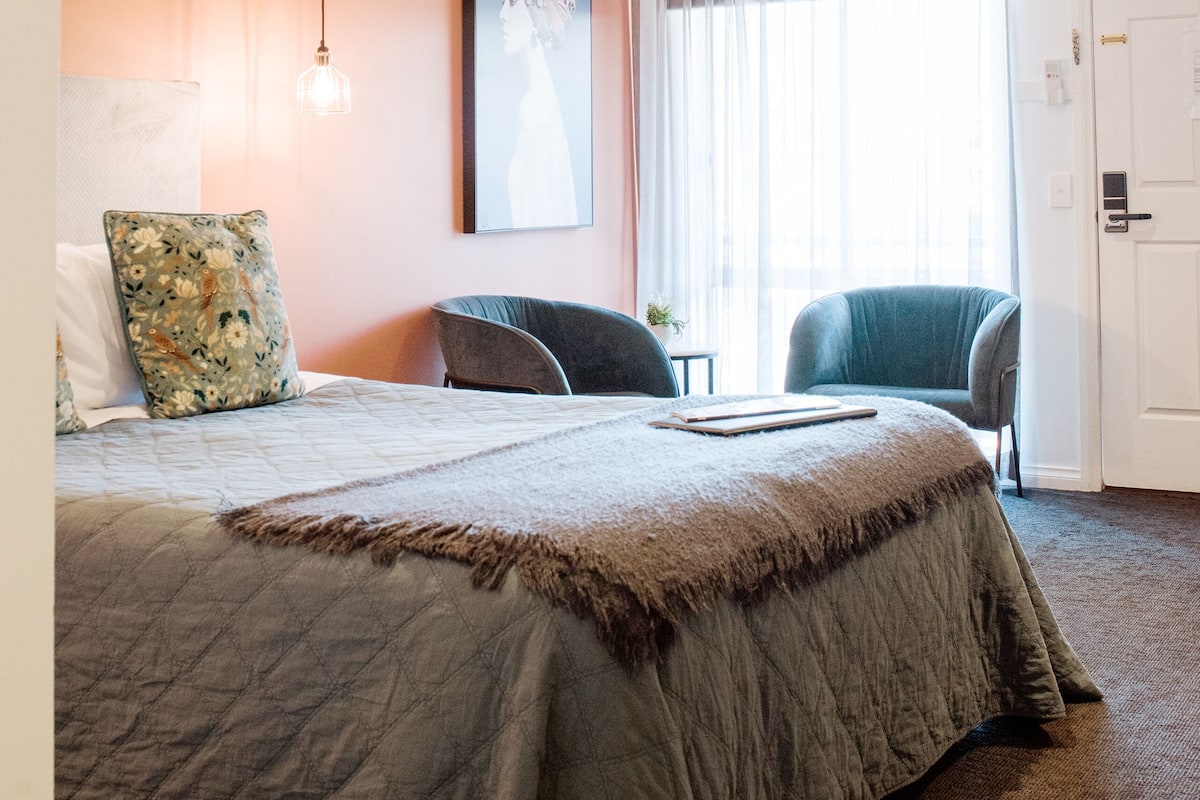 A hotel room with a large bed, two chairs, a painting on the wall, and a window with curtains.