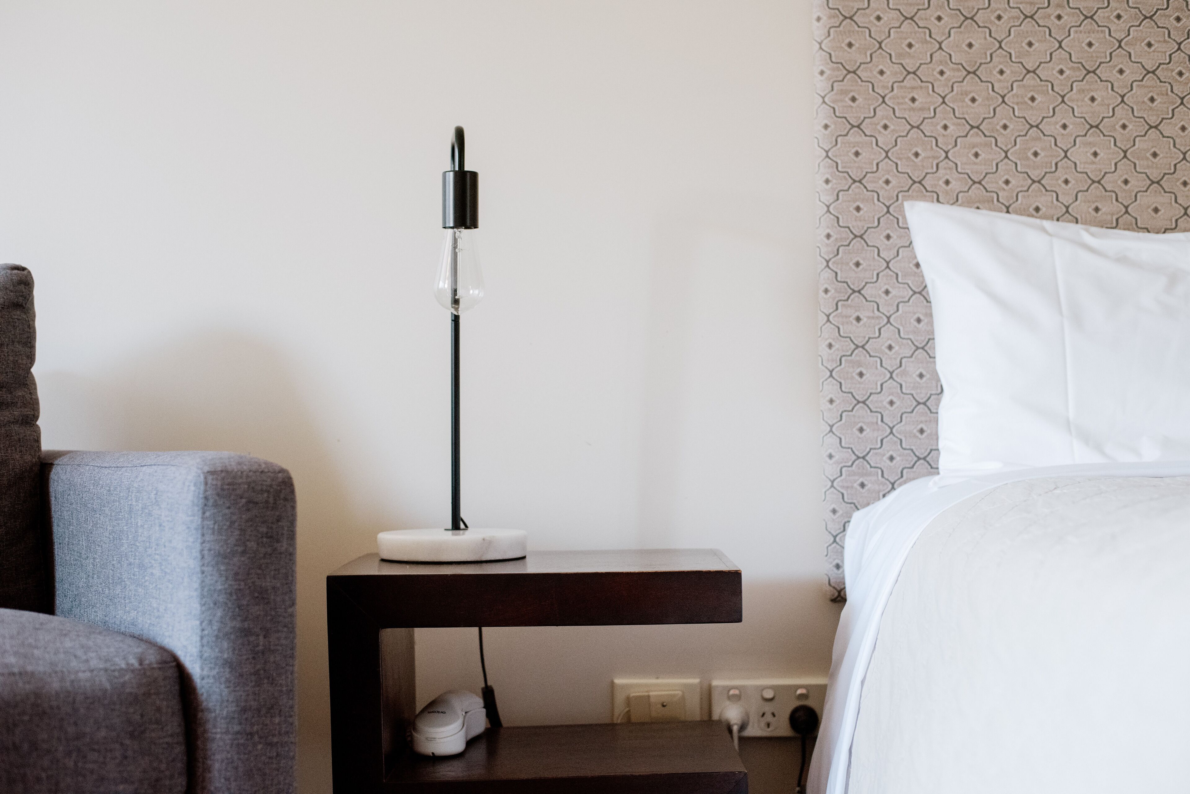 Un cuarto de hotel ordenado con cama, un sillón gris, una mesita de noche con lámpara y una pared con estampado.