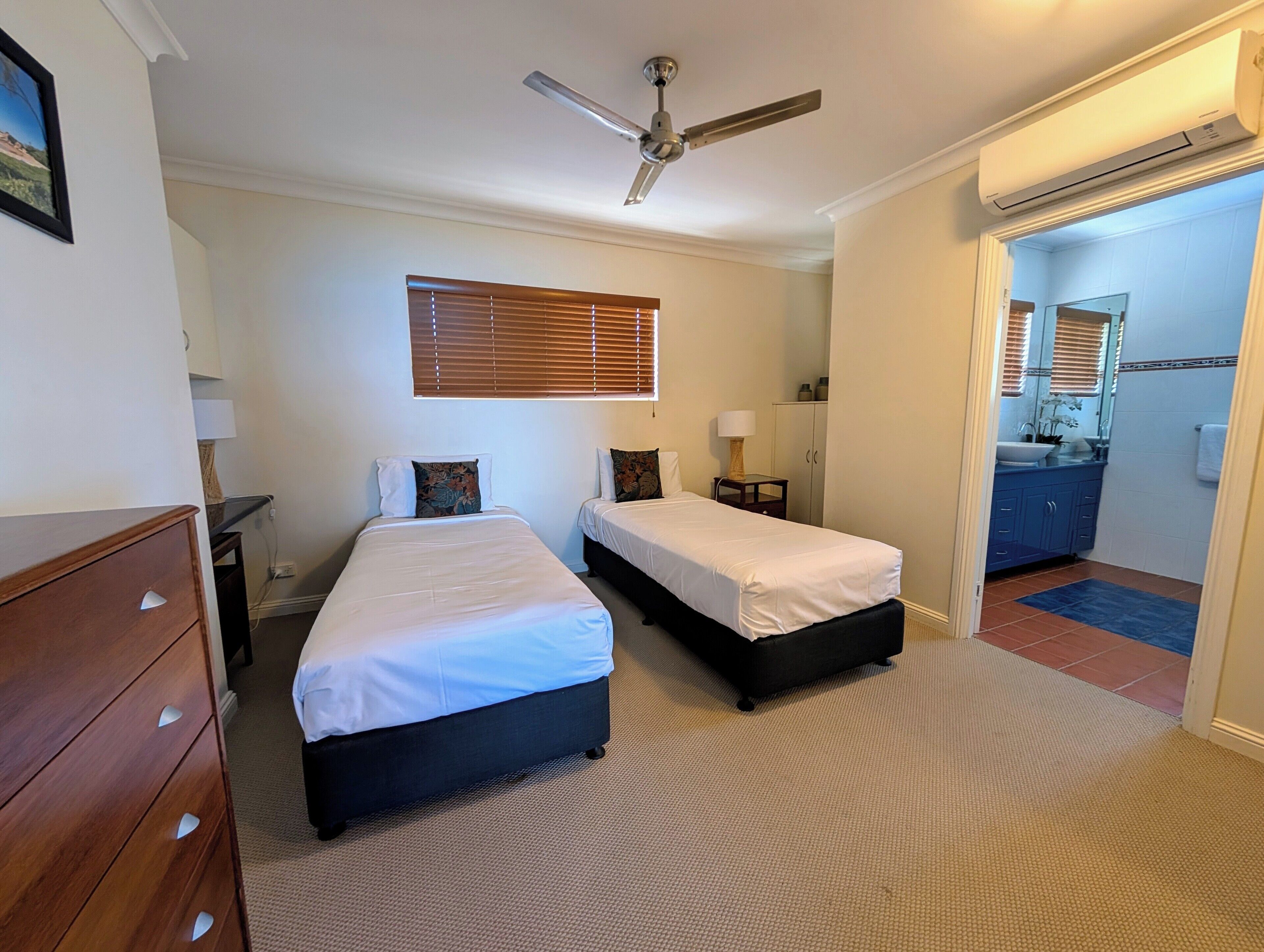 Une chambre avec deux lits, un ventilateur de plafond, une commode, une table de chevet et une salle de bain visible à travers une porte ouverte.
