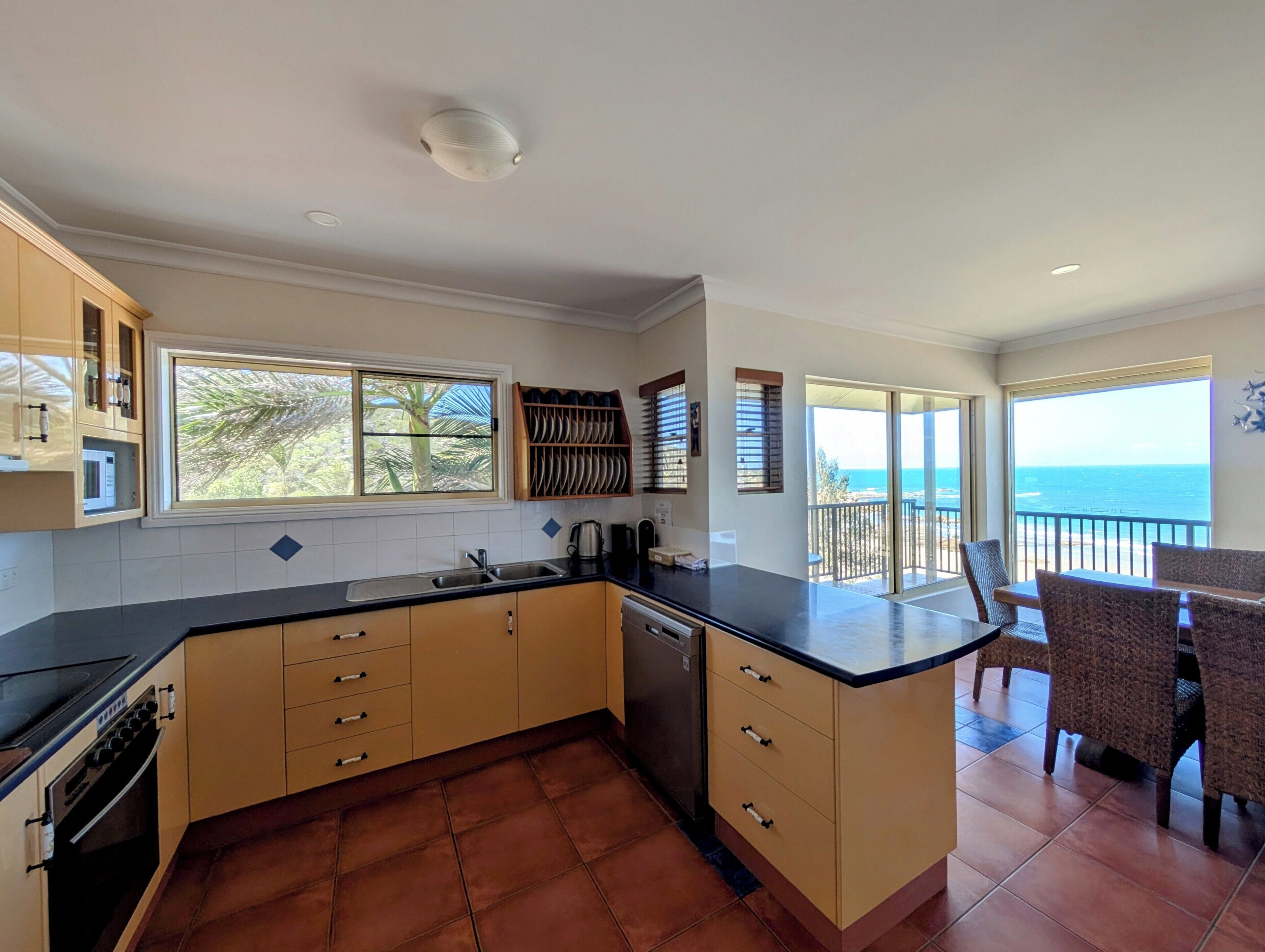 A kitchen with a view of the ocean, a dining area with chairs, and a balcony with a railing.