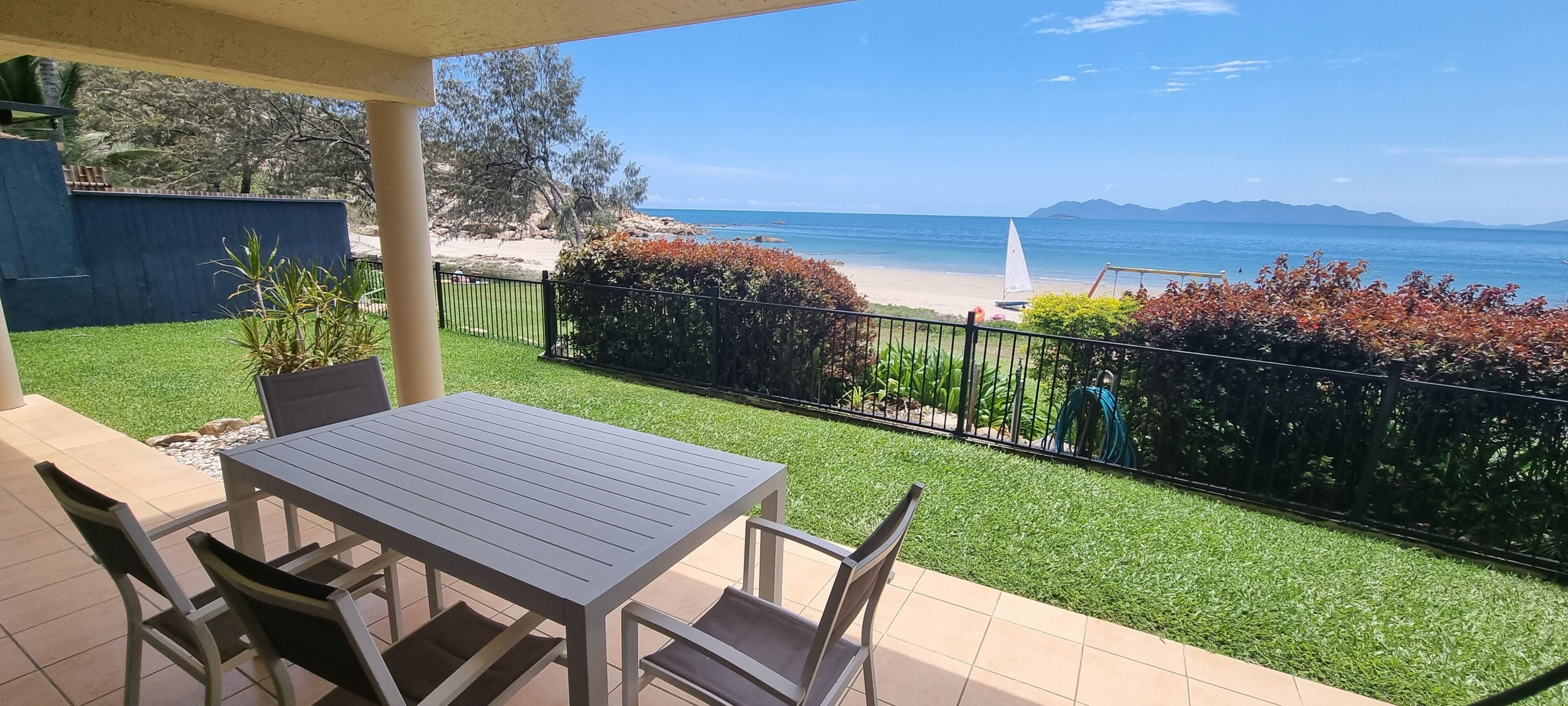 Eine Terrasse mit Tisch und Stühlen, Blick auf den Strand und ein Segelboot in der Ferne.