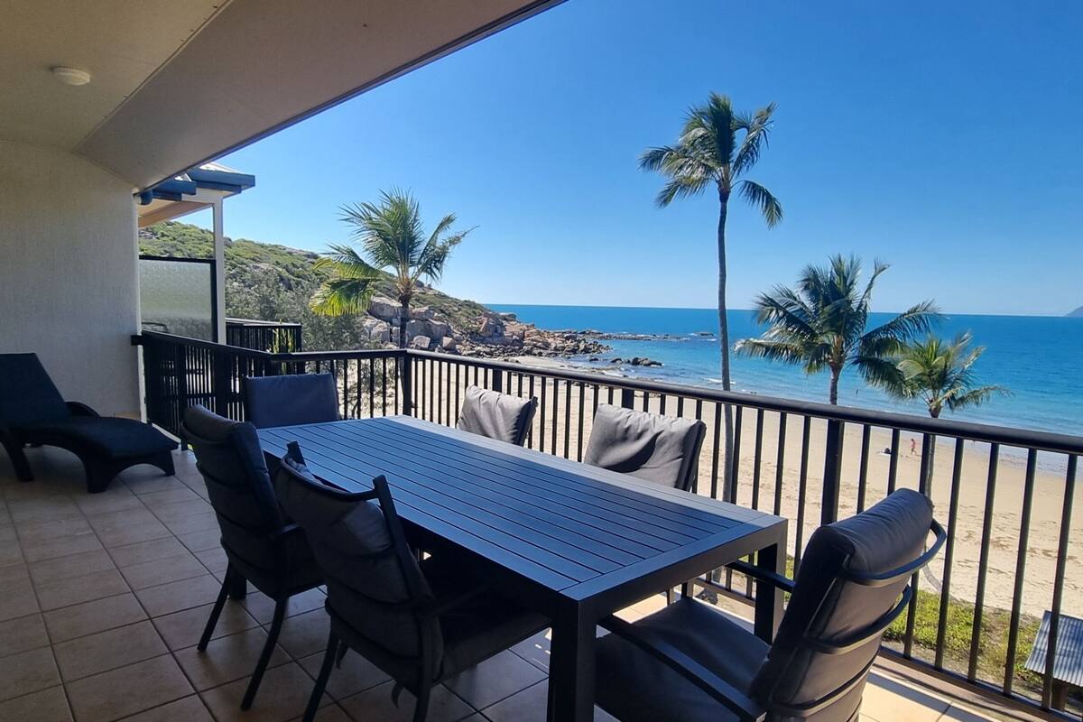 A balcony with a table and chairs, overlooking a beach with palm trees and the ocean.