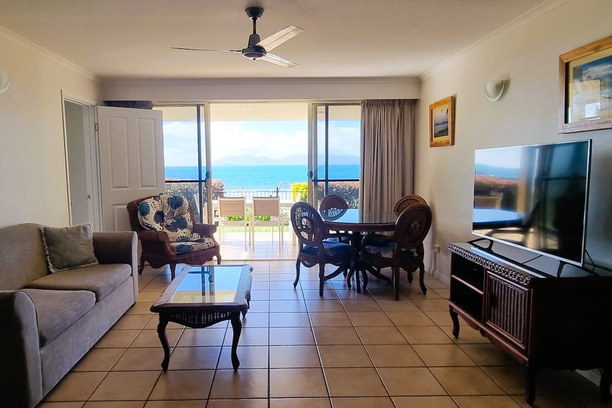 A living room with a sofa, coffee table, TV, and a balcony with a view of the sea.