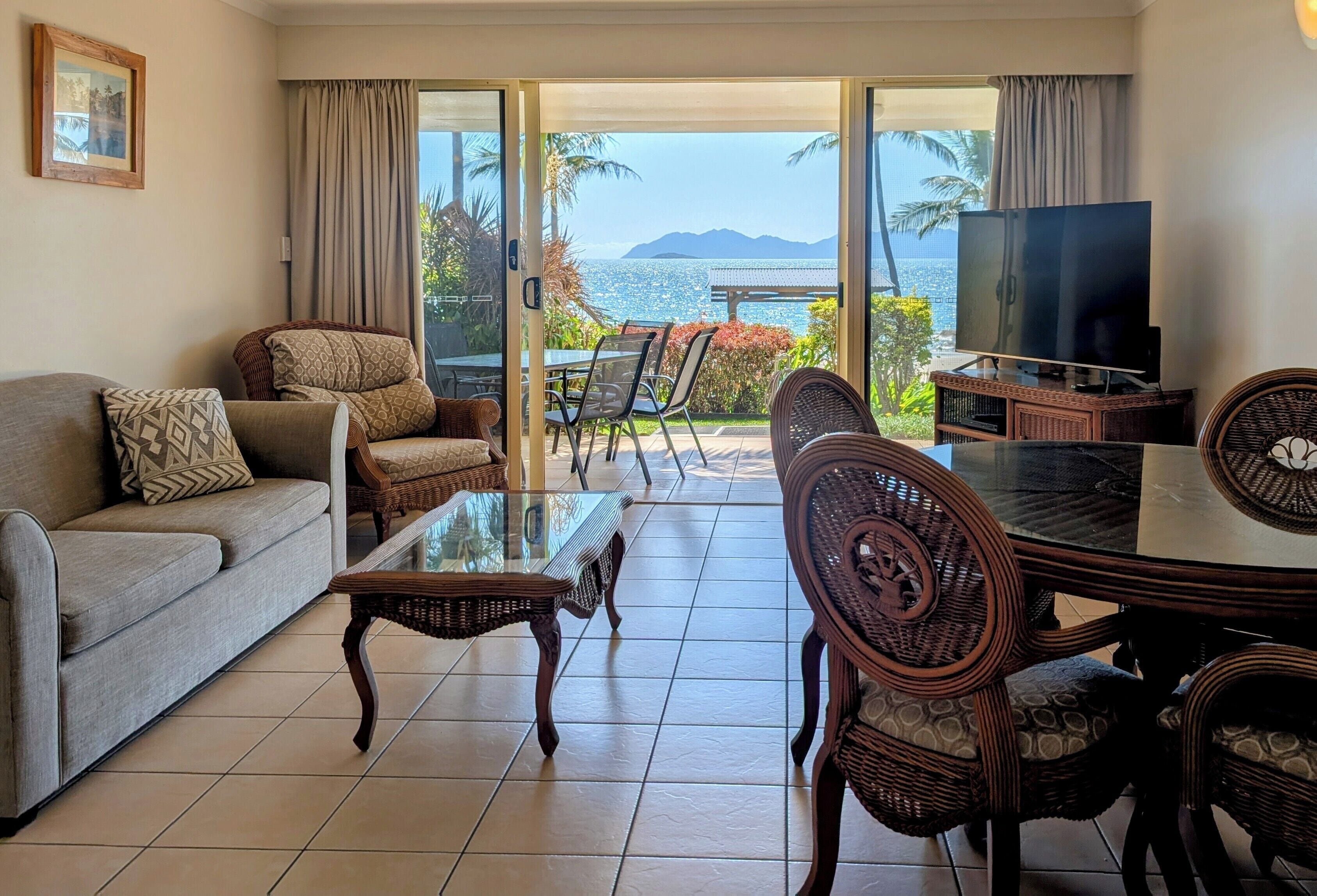 A living room with a sofa, chairs, a glass table, a TV, and a view of the ocean.