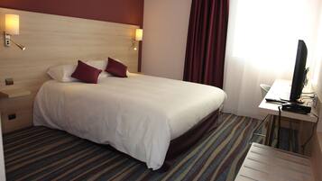 A hotel room with a large bed, two bedside lamps, a desk with a computer, and a window with curtains.