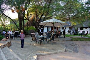 Un patio al aire libre con suelo de piedra, mesas y sillas. Se ve gente cenando y una persona de pie cerca de la entrada.