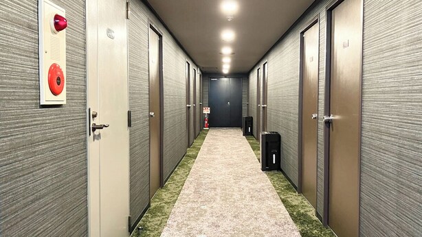 A hallway with a carpeted floor, doors on both sides, and a fire alarm on the left. - HOTEL LiVEMAX Chofu-Ekimae (Chofu)