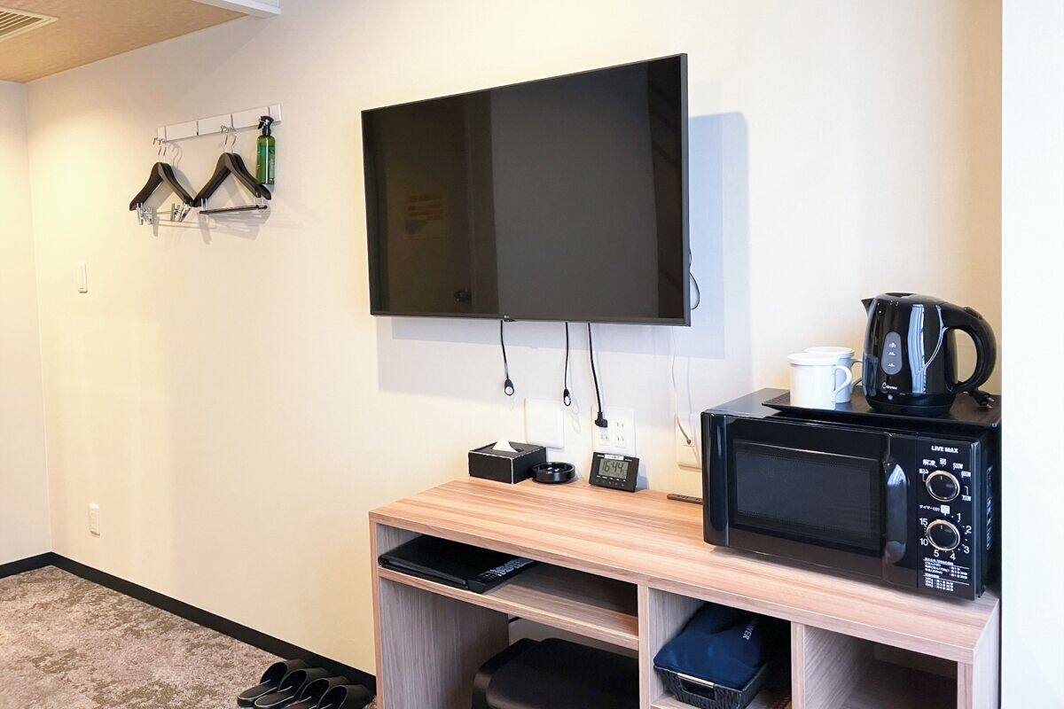 A compact room with a flat-screen TV mounted on the wall, a small wooden desk with a microwave, and a shoe rack against the wall.