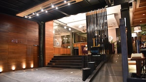 A modern hotel lobby with a wooden reception desk, a staircase leading to upper floors, and a glass partition separating the lobby from the dining area.