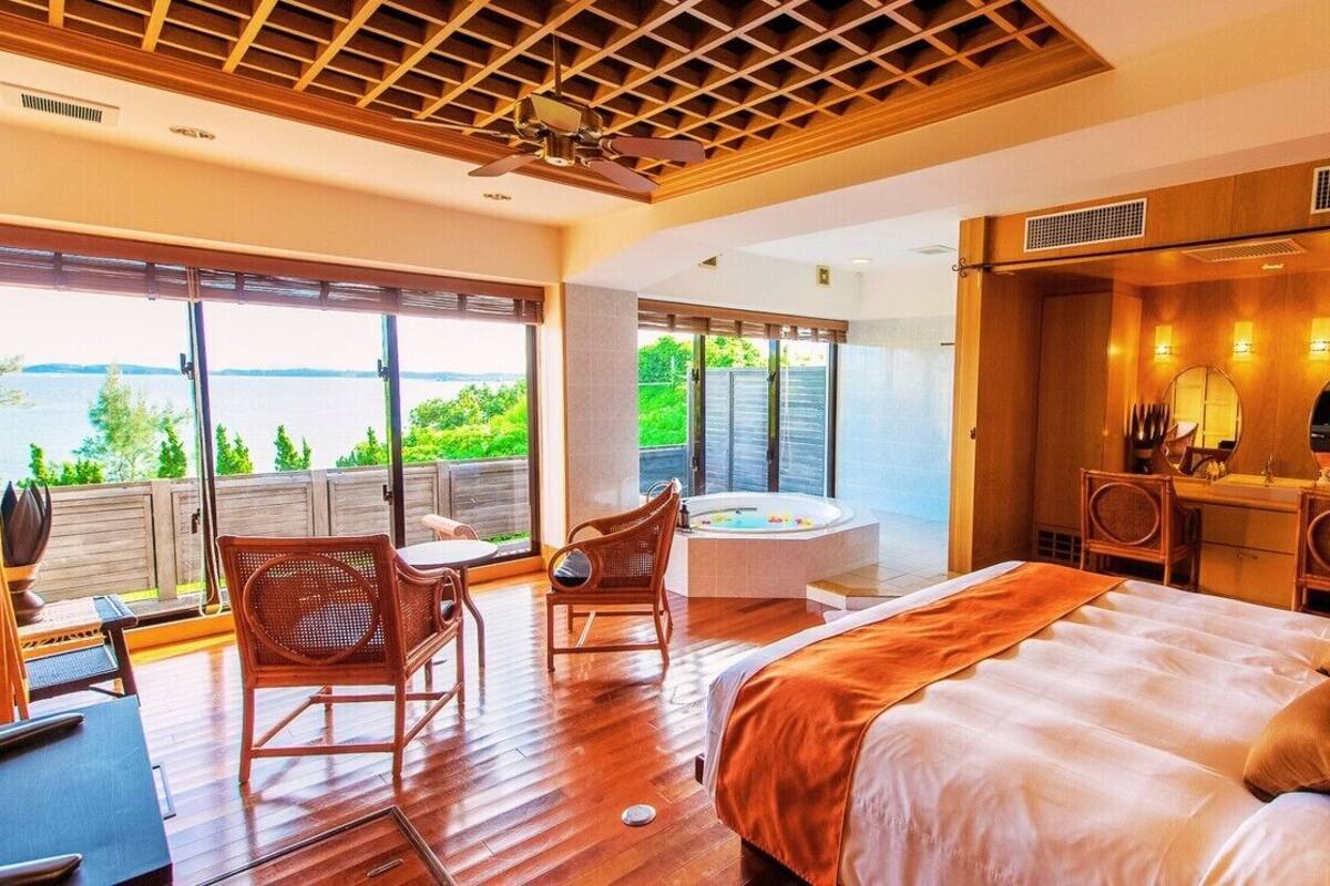 A bedroom with a large bed, a sitting area with chairs, a bathtub, and a view of the water.