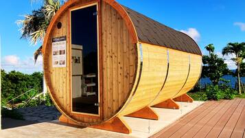 A wooden sauna with a curved roof, situated on a wooden deck with a view of the sea.