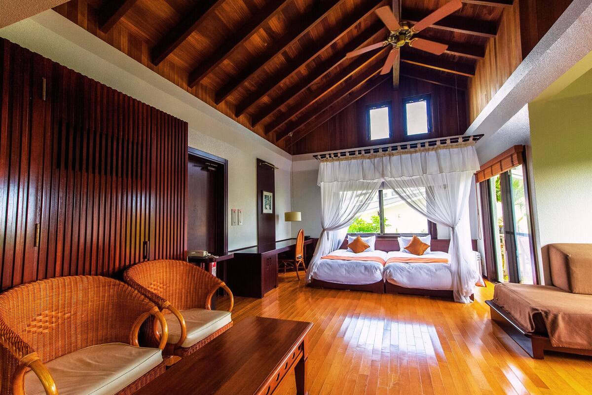A spacious room with a high ceiling, wooden beams, a bed with a canopy, wicker furniture, and a wooden floor.