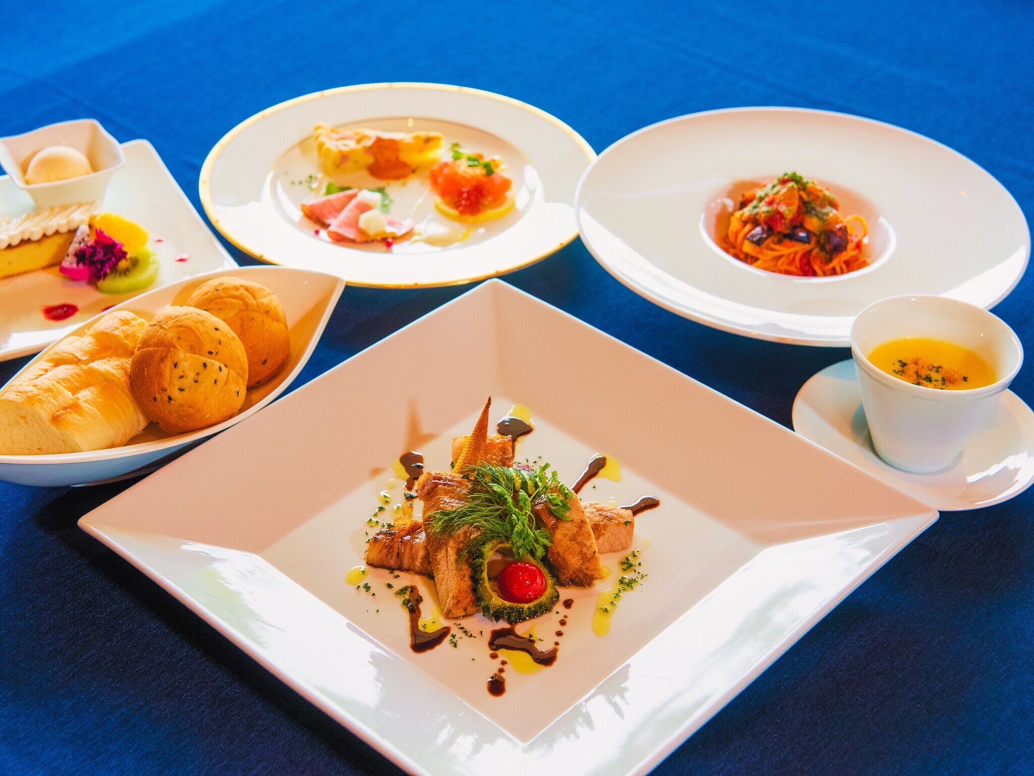 A table with various dishes, including a square plate with a fish dish, a round plate with a salad, and a small bowl with a soup.