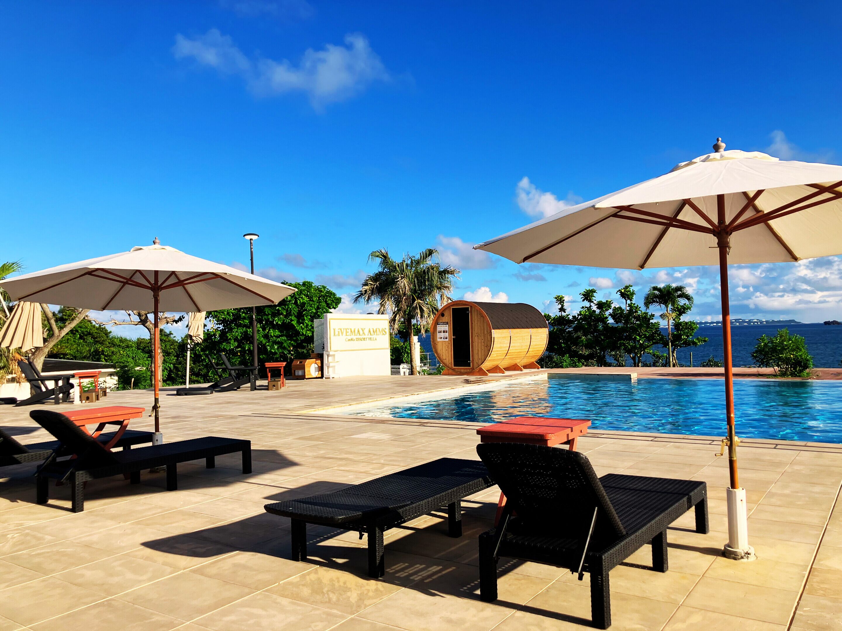 A poolside area with lounge chairs, umbrellas, and a wooden barrel sauna.