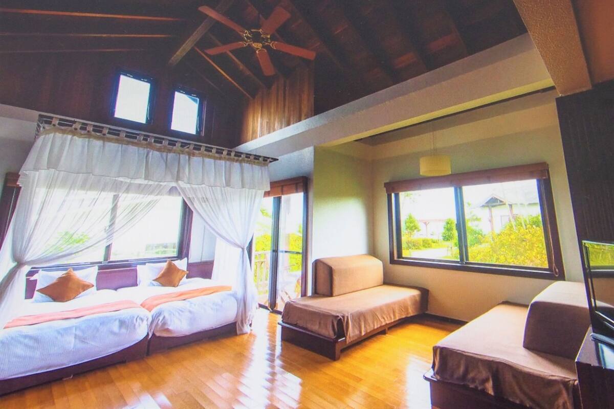 A room with a bed, a sofa, and a desk. There are windows and a ceiling fan.