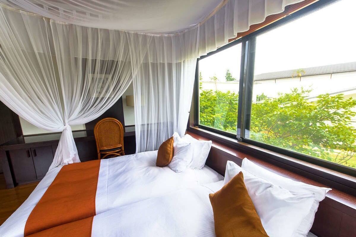 A hotel room with a bed, white curtains, a view of greenery outside, and a wooden chair.