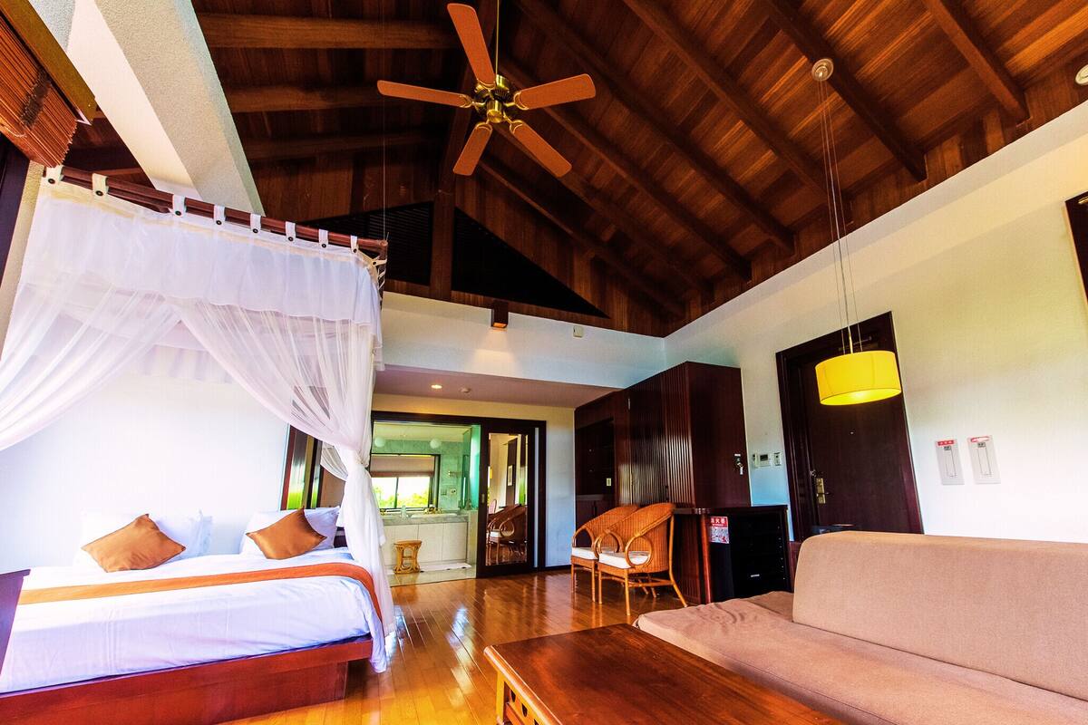 A spacious room with a bed, a sofa, a wooden table, and a ceiling fan.