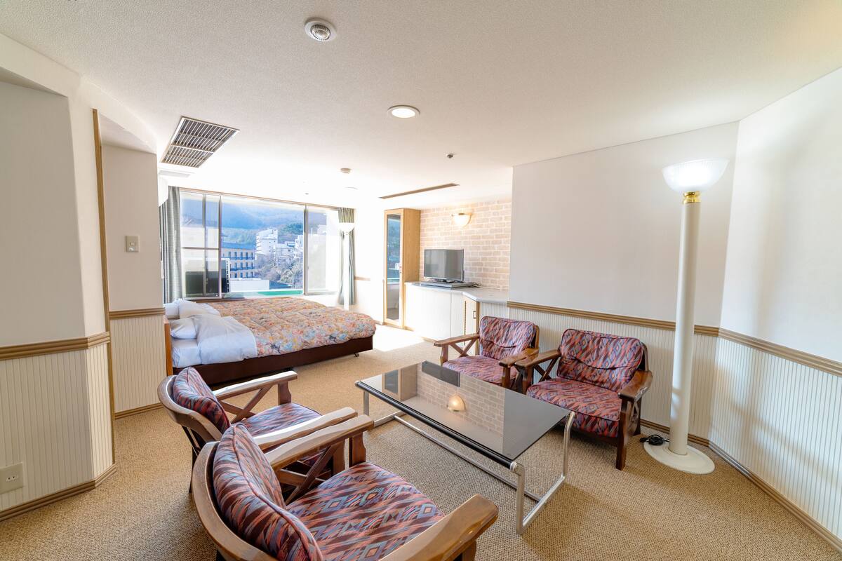 Une chambre d’hôtel avec un lit, une table à plateau en verre, des chaises à motifs, un canapé, une télévision et une vue sur les immeubles à travers de grandes fenêtres.