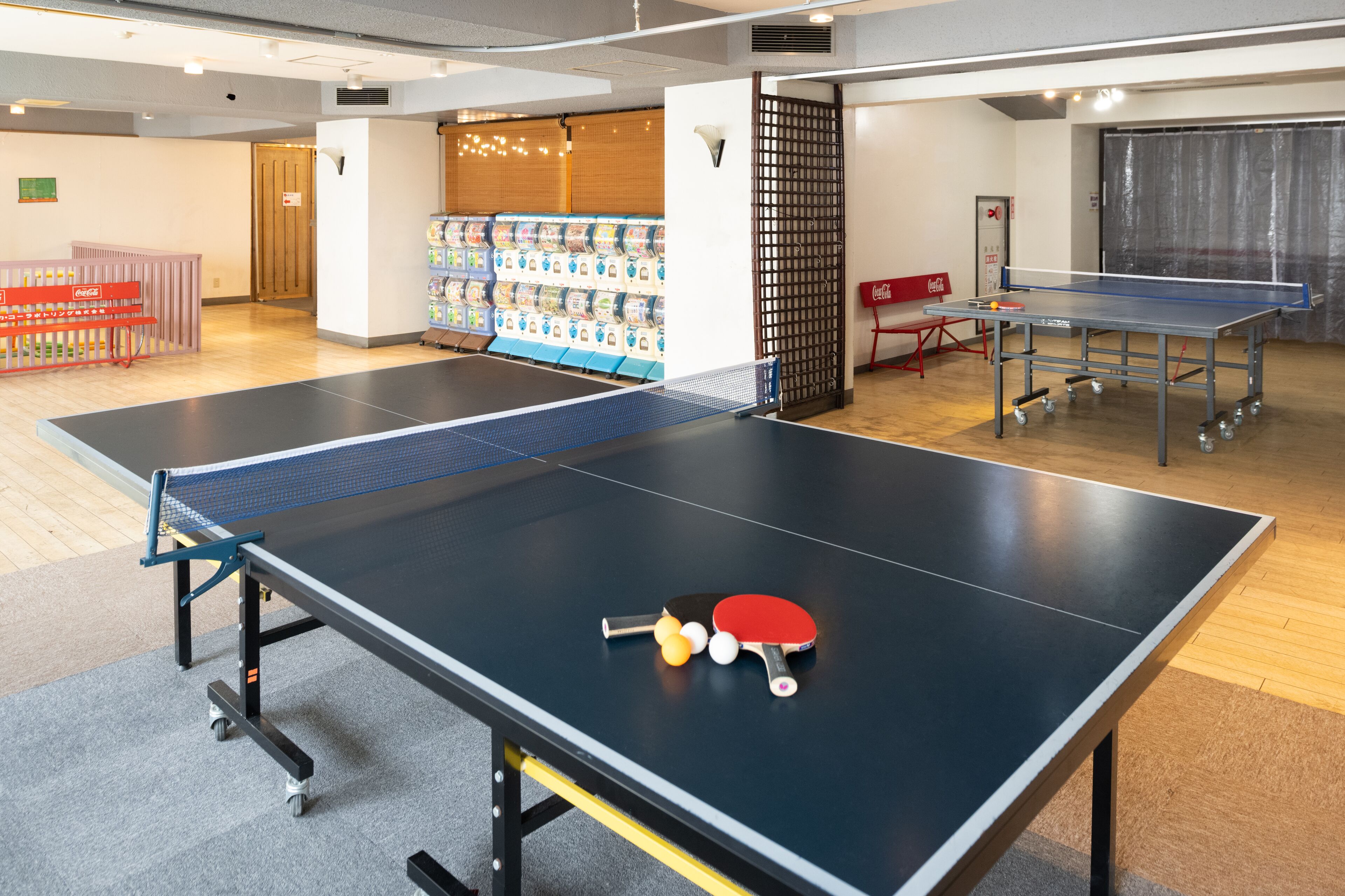 Une table de ping-pong avec un filet, deux raquettes et trois balles, dans une pièce dont le sol est en bois, qui contient également un distributeur automatique et un réfrigérateur à boissons Coca-Cola.