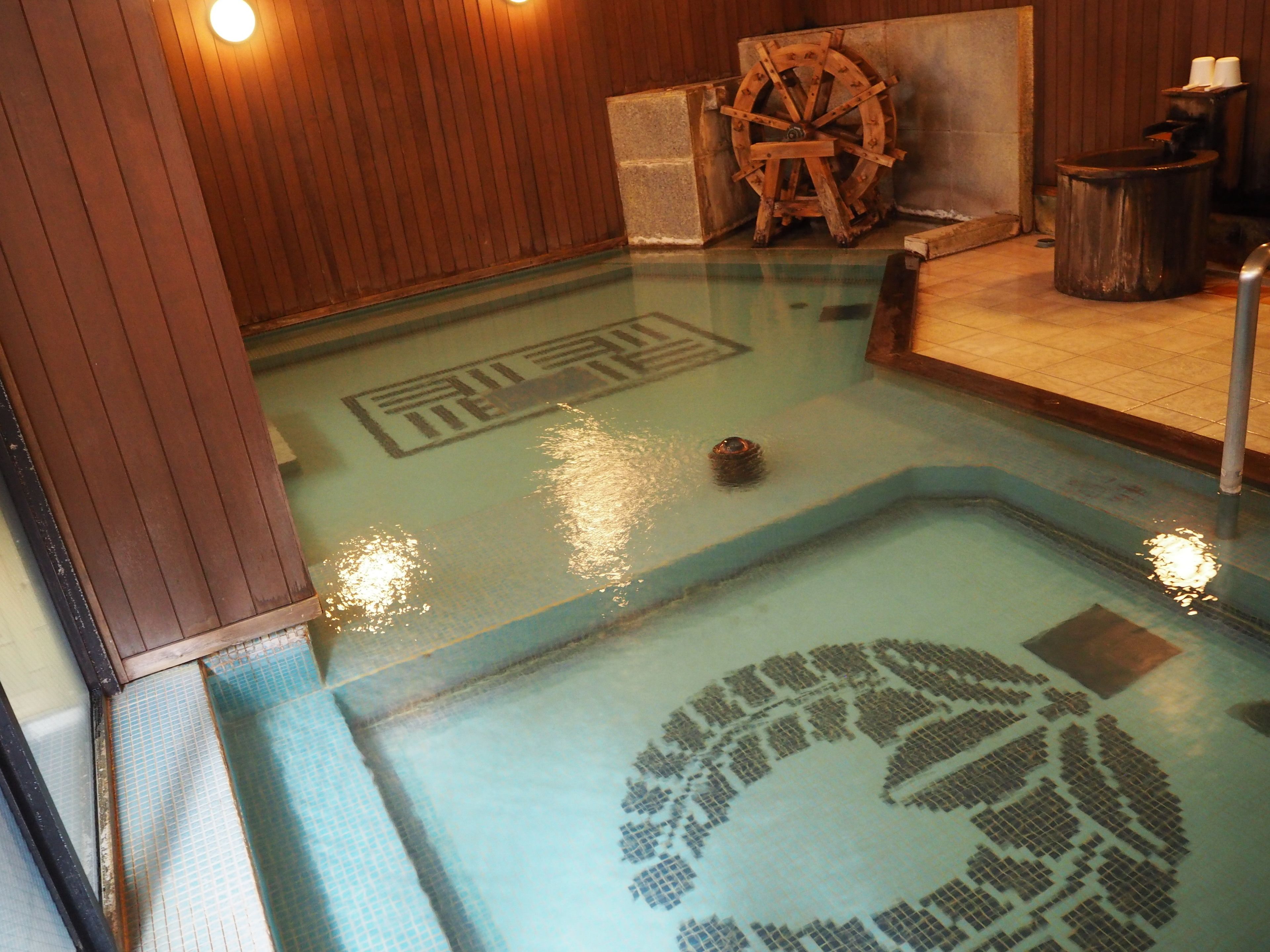A traditional Japanese onsen with a wooden interior, a stone basin, and a wooden wheel.