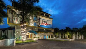 Een hotel met een groot bord waarop 'Surfer Hotel' staat, en een goed verlichte entree met een palmboom en een decoratieve lichtinstallatie.