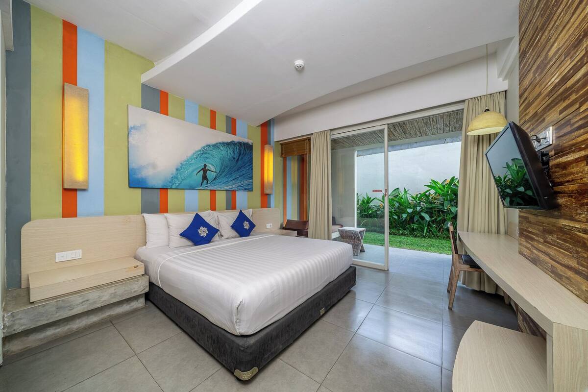 Een moderne hotelkamer met een groot bed, een bureau en een glazen schuifdeur naar een balkon met planten.