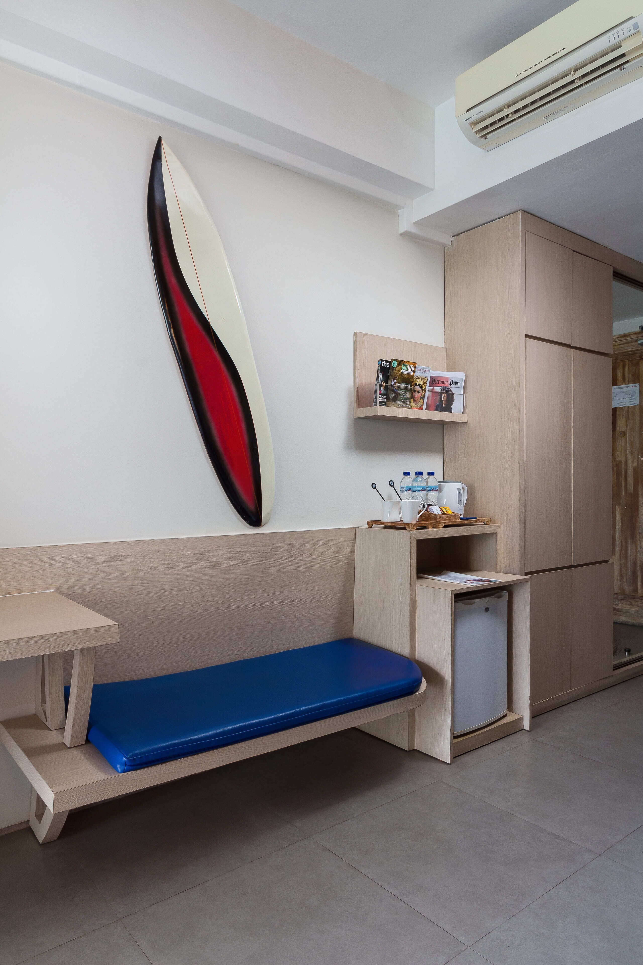 Ein kleines Zimmer mit einer hölzernen Sitzbank, einem kleinen Tisch, einem an der Wand befestigten Surfbrett, einem Regal mit Zeitschriften und einem Kühlschrank.