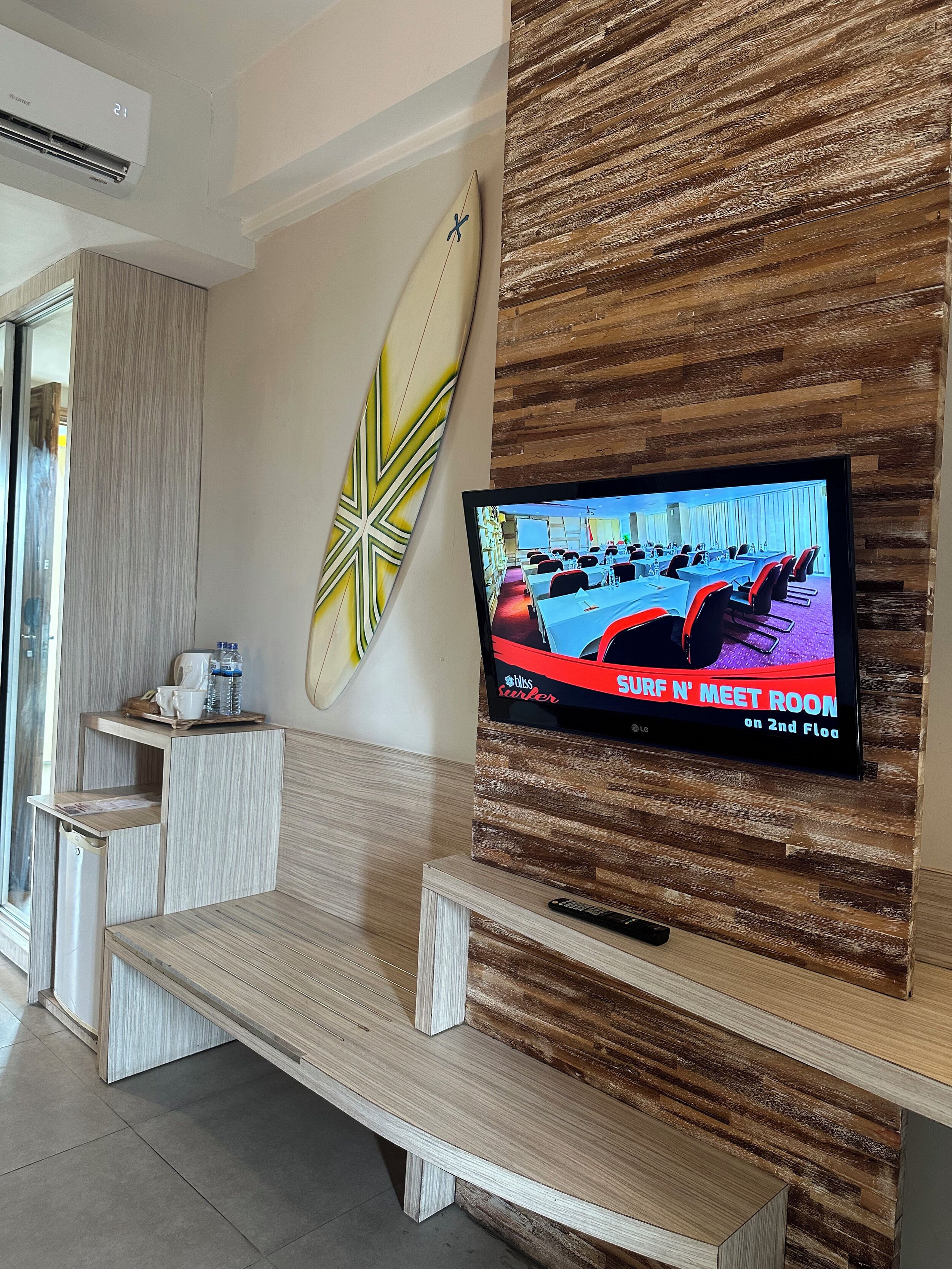 Una habitación de hotel moderna con un televisor de pantalla plana montado en una pared de madera, una decoración con una tabla de surf y una zona de estar con un banco de madera.
