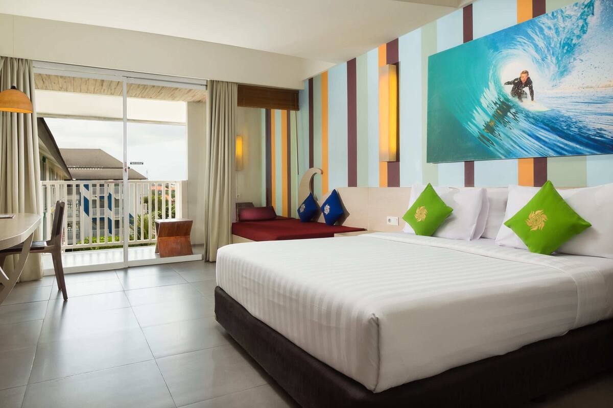 Een hotelkamer met een bed, een bureau, een stoel en een balkon met uitzicht op gebouwen.