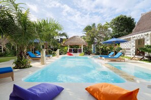 A swimming pool with lounge chairs and umbrellas, surrounded by greenery and a thatched-roof structure. - Hotel Puri Tempo Doeloe (Denpasar)