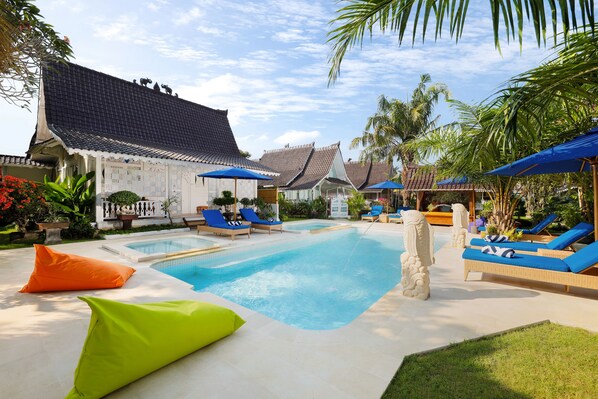 A swimming pool with lounge chairs and umbrellas, surrounded by tropical plants and traditional architecture. - Hotel Puri Tempo Doeloe (Denpasar)